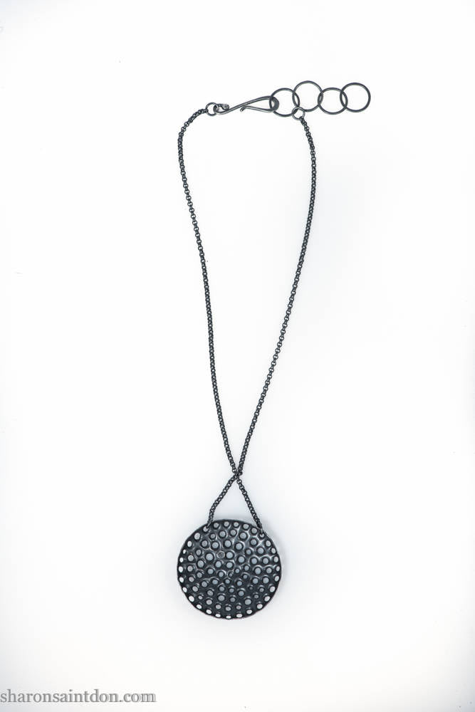 Large round, handcrafted sterling silver pendant necklace with perforated holes and oxidized black finish, accented with cubic zirconia, on black chain.