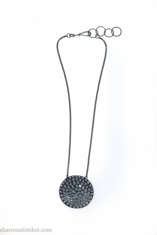 Large round, handcrafted sterling silver pendant necklace with perforated holes and oxidized black finish, accented with cubic zirconia, on black chain.