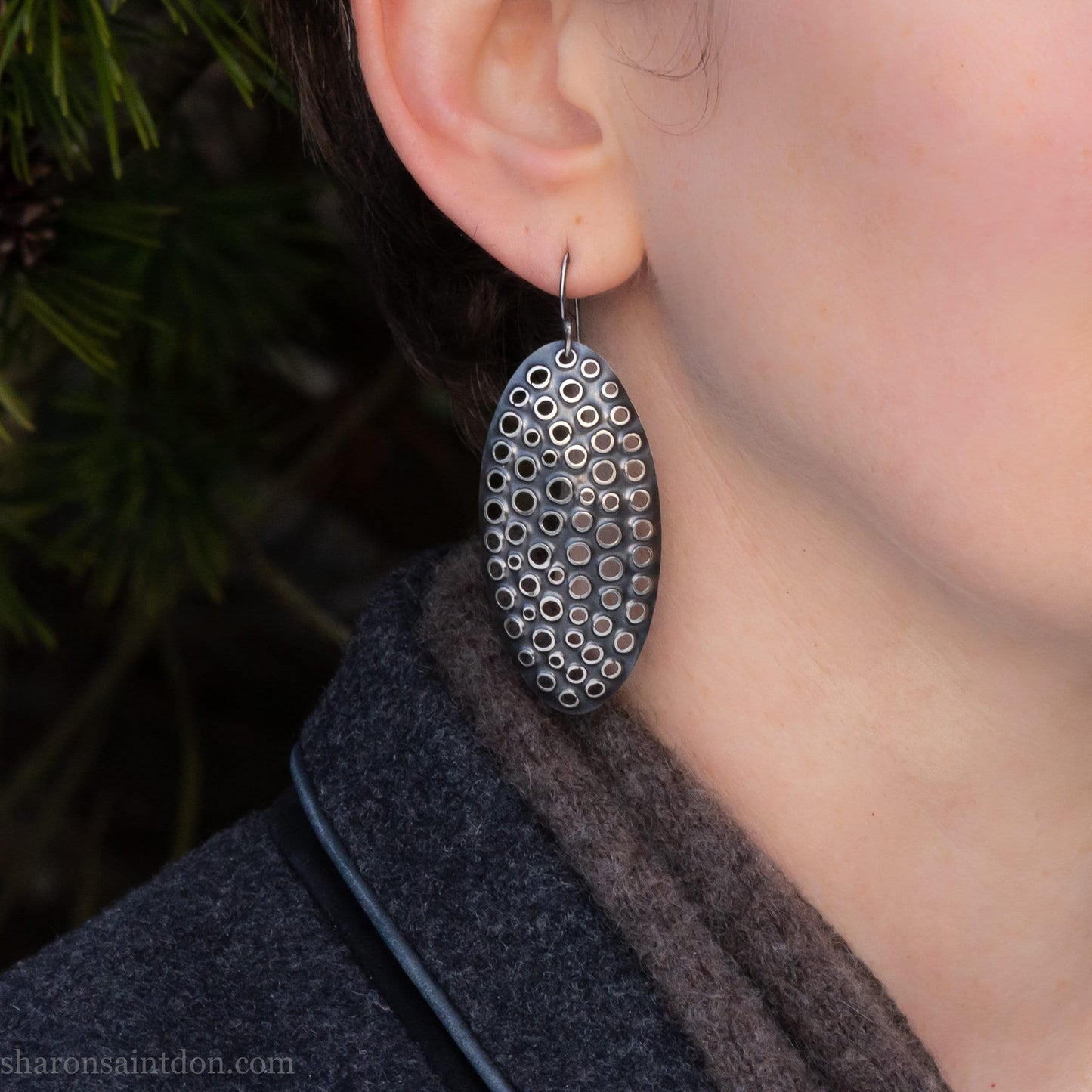 Large oval sterling silver dangle earrings with hand-punched detailed holes and antiqued finish, worn on ear.