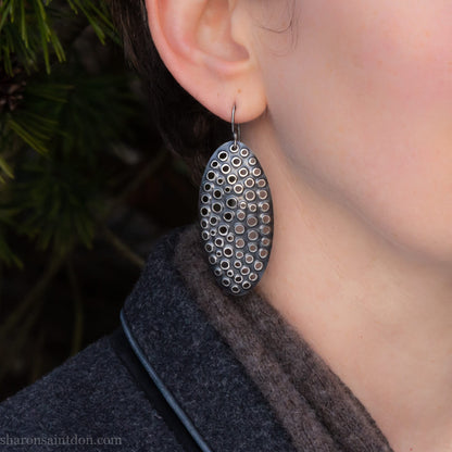Large oval sterling silver dangle earrings with hand-punched detailed holes and antiqued finish, worn on ear.