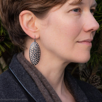 Large oval sterling silver dangle earrings with hand-punched detailed holes and antiqued finish, worn on ear.