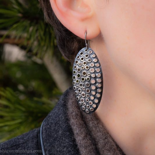 Large oval sterling silver dangle earrings with hand-punched detailed holes and antiqued finish, worn on ear.