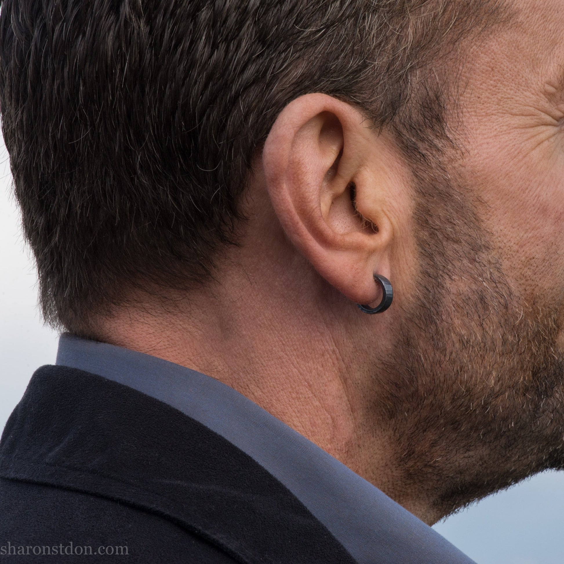 Close up photo of a man wearing handcrafted oxidized black 14mm x 4mm hammered solid sterling silver hoop earrings.
