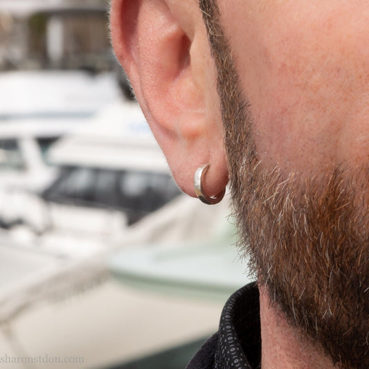 Handmade solid sterling silver hoop earrings, hammered by hand with a matte, brushed, shiny finish. SIZE: 12mm exterior diameter, 9mm interior, 2mm wide. and 1.5mm thick. Pictured on a man with light skin and brown beard.