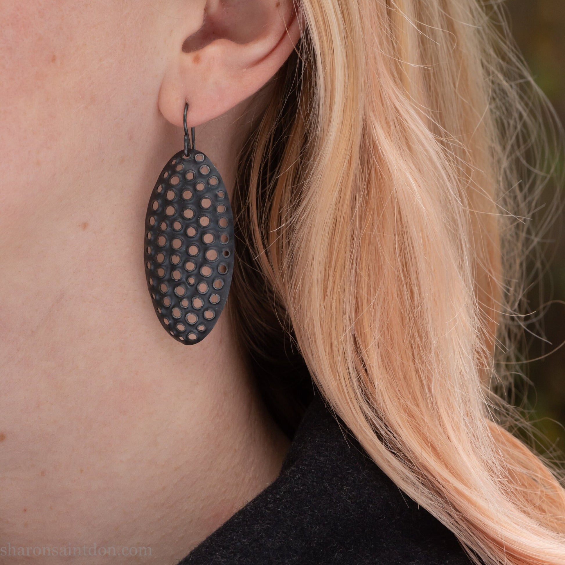 Woman wearing oxidized silver dangle earrings with hand-punched oval apertures, 50mm long.