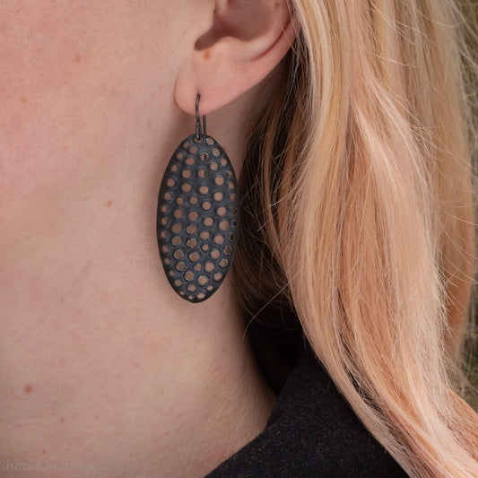 Woman wearing oxidized silver dangle earrings with hand-punched oval apertures, 50mm long.