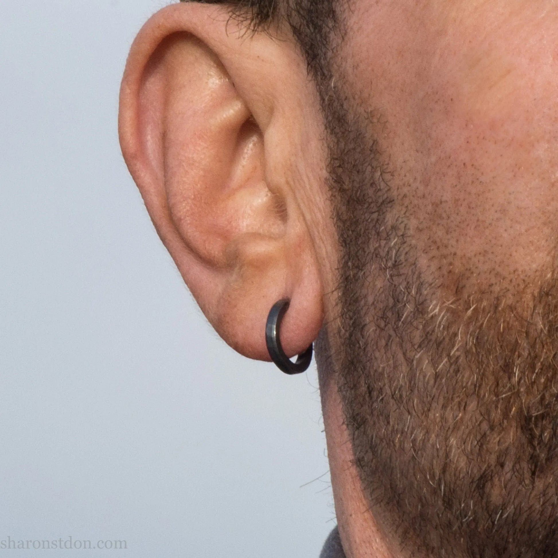 Small black hammered sterling silver hoop earrings worn by man, side profile, 14x2mm.